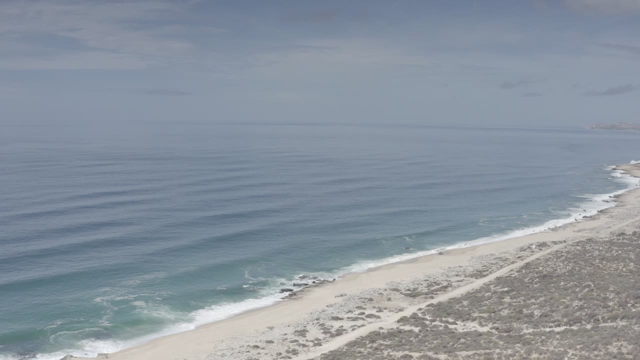 The Los Cabos shore runs diagonally across the lower frame as soft gray-blue water forms parallel wave lines, with no visible people on the beach.