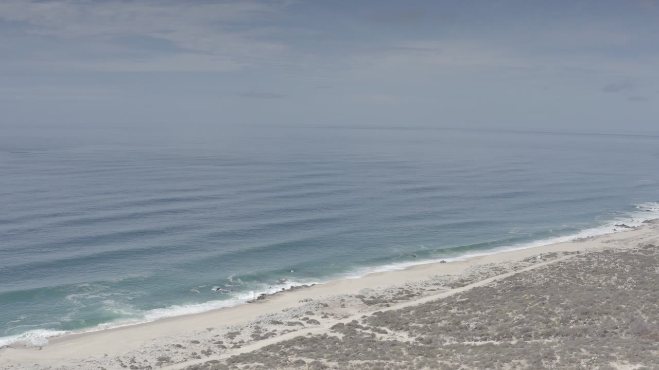 Aerial view over the Los Cabos coast with muted ocean bands, a pale sandy beach, scrubby foreground dunes, and no visible people.
