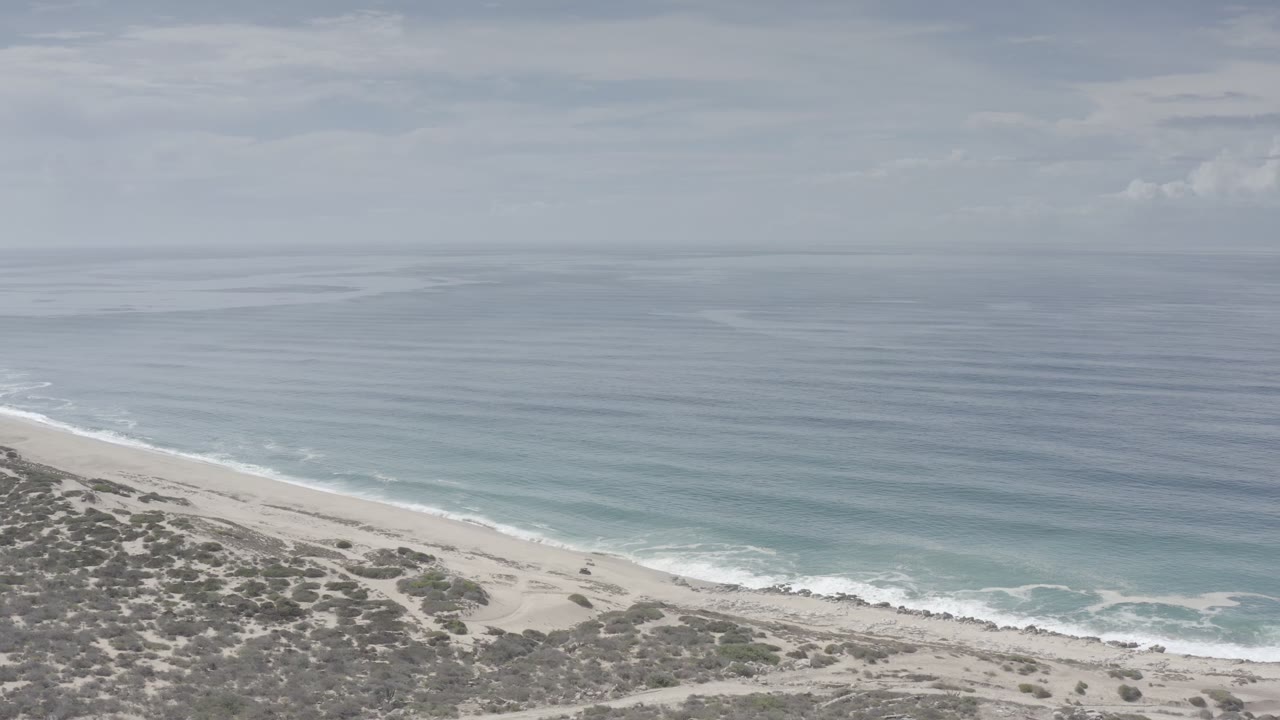 Opening frame of a Los Cabos, Mexico beach overlook with scrub dunes in the foreground, pale sand below, blue ocean bands beyond, and no visible people.
