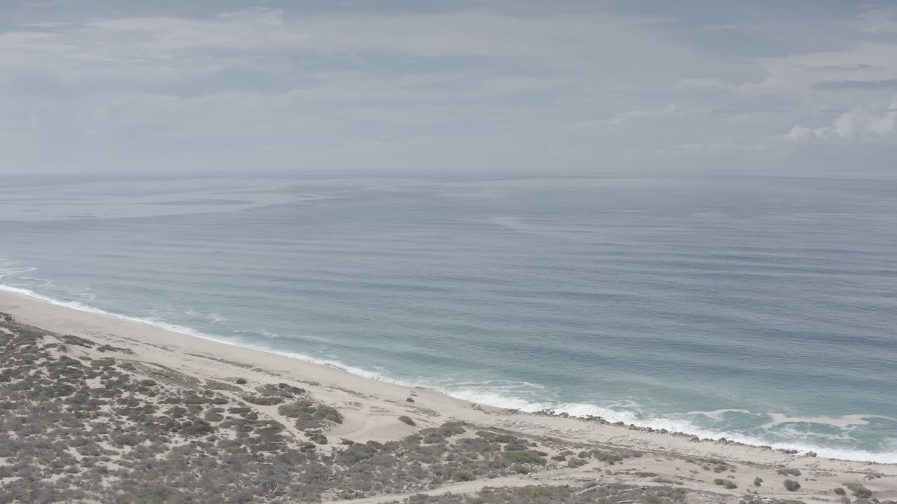Los Cabos coastline frame with a diagonal strip of pale beach, low scrub in the lower left, calm blue water, and no visible people.