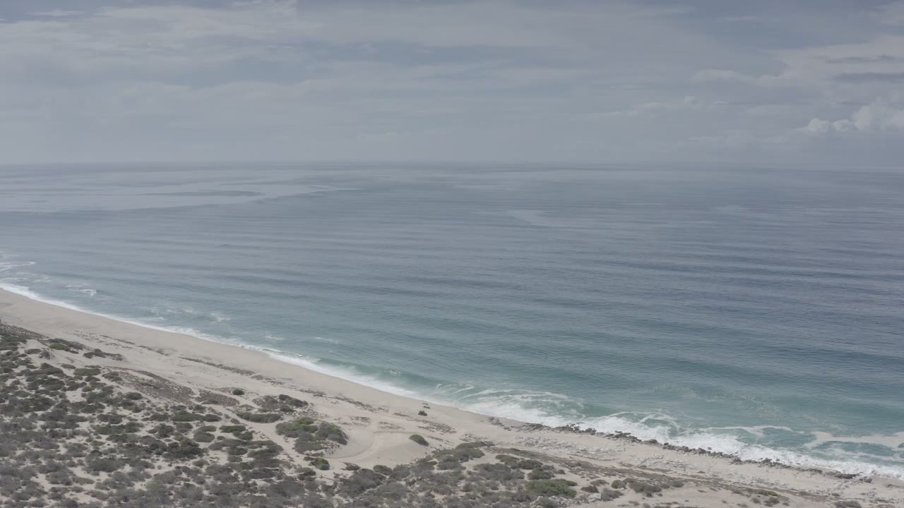 Aerial view in Los Cabos, Mexico, with the shoreline angled across the lower frame, small white waves, open sea, and no visible people.