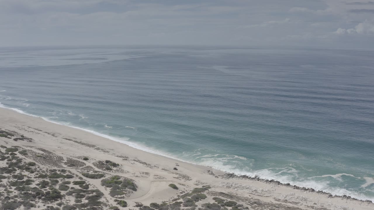 Opening aerial frame over a Los Cabos beach, with dune scrub at the lower left, pale sand crossing diagonally, blue-green water beyond, and no visible people.