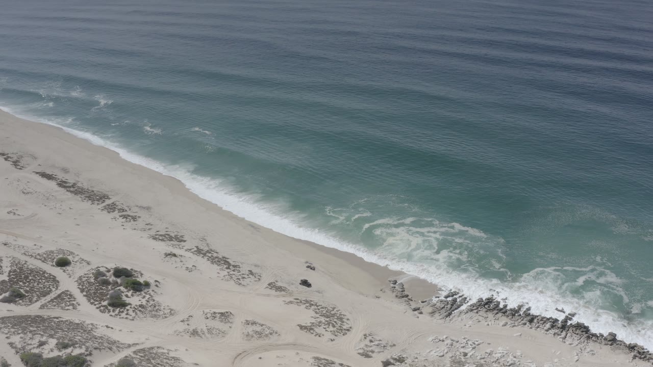 Opening aerial frame of Los Cabos shoreline with blue green ocean meeting pale sand, white foam at the waterline, scattered rocks, scrub patches, and no visible people.