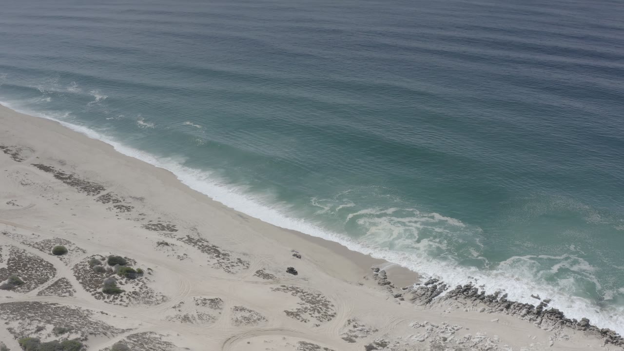 Aerial Los Cabos beach frame with a wider patch of foamy surf curling onto the sand near the rocky lower shoreline, with no visible people.