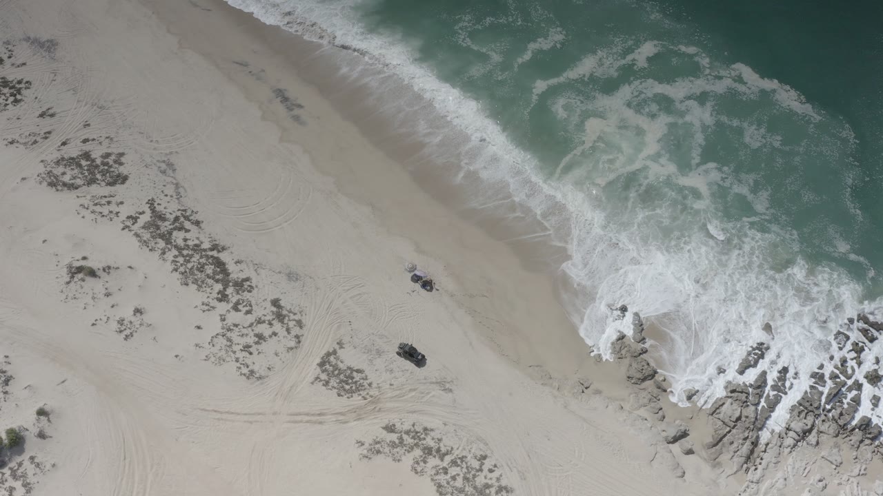 Overhead view of a Los Cabos beach at 0:00 with teal surf at the upper right, pale sand, shoreline rocks, track marks, a small dark object on the sand, and no visible people.