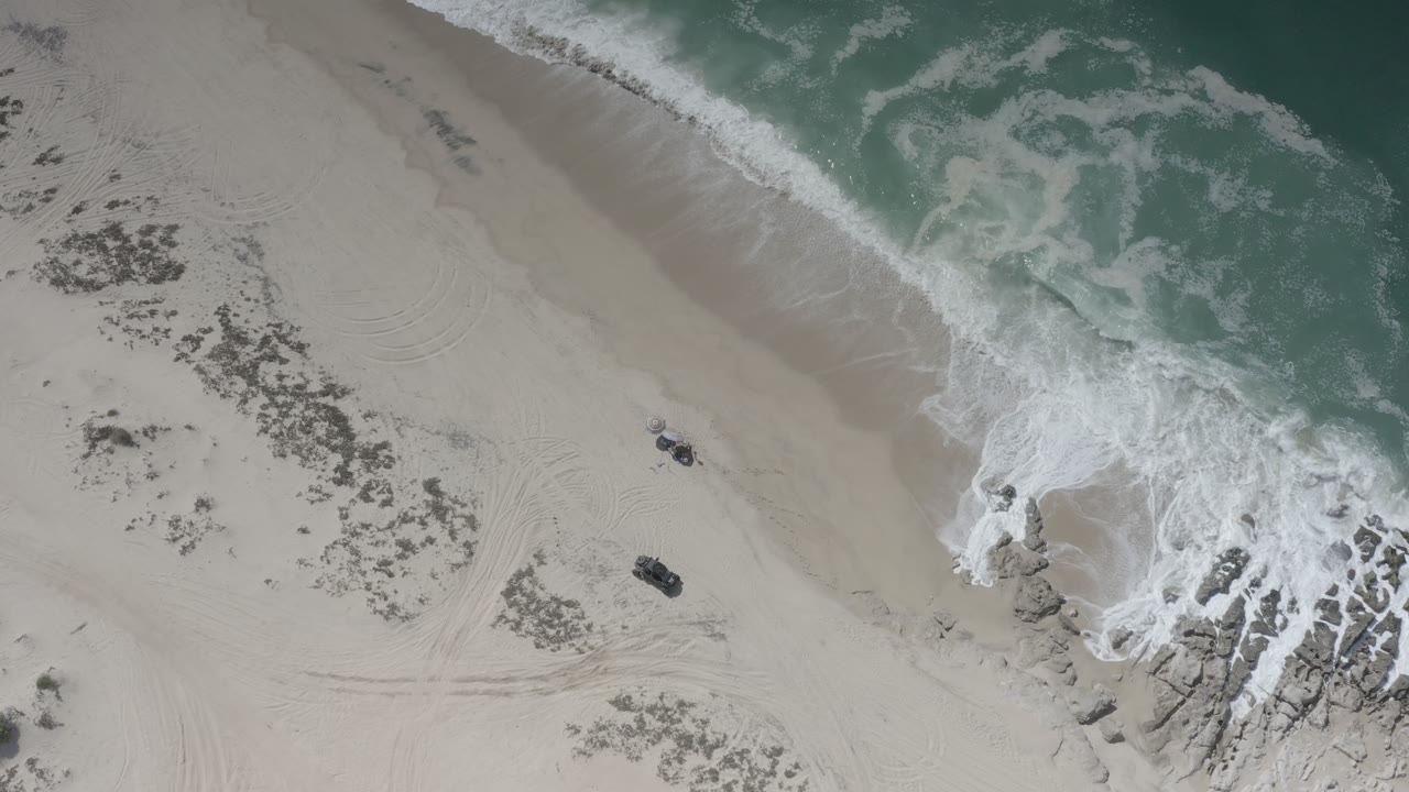 Top-down beach view with turquoise water, breaking foam, rocks, and dark sand object.