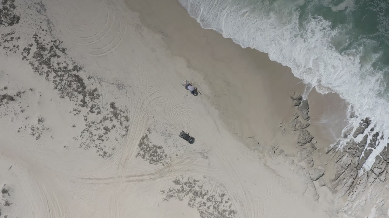 Overhead Los Cabos beach frame with white surf at the top edge, pale sand across the center, rocks on the right, faint track marks, and no visible people.