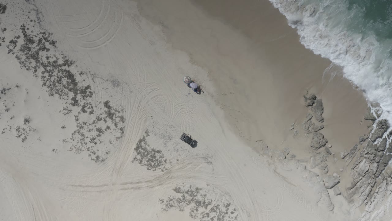 Overhead beach with surf, rocks, two people, and a small dark vehicle.