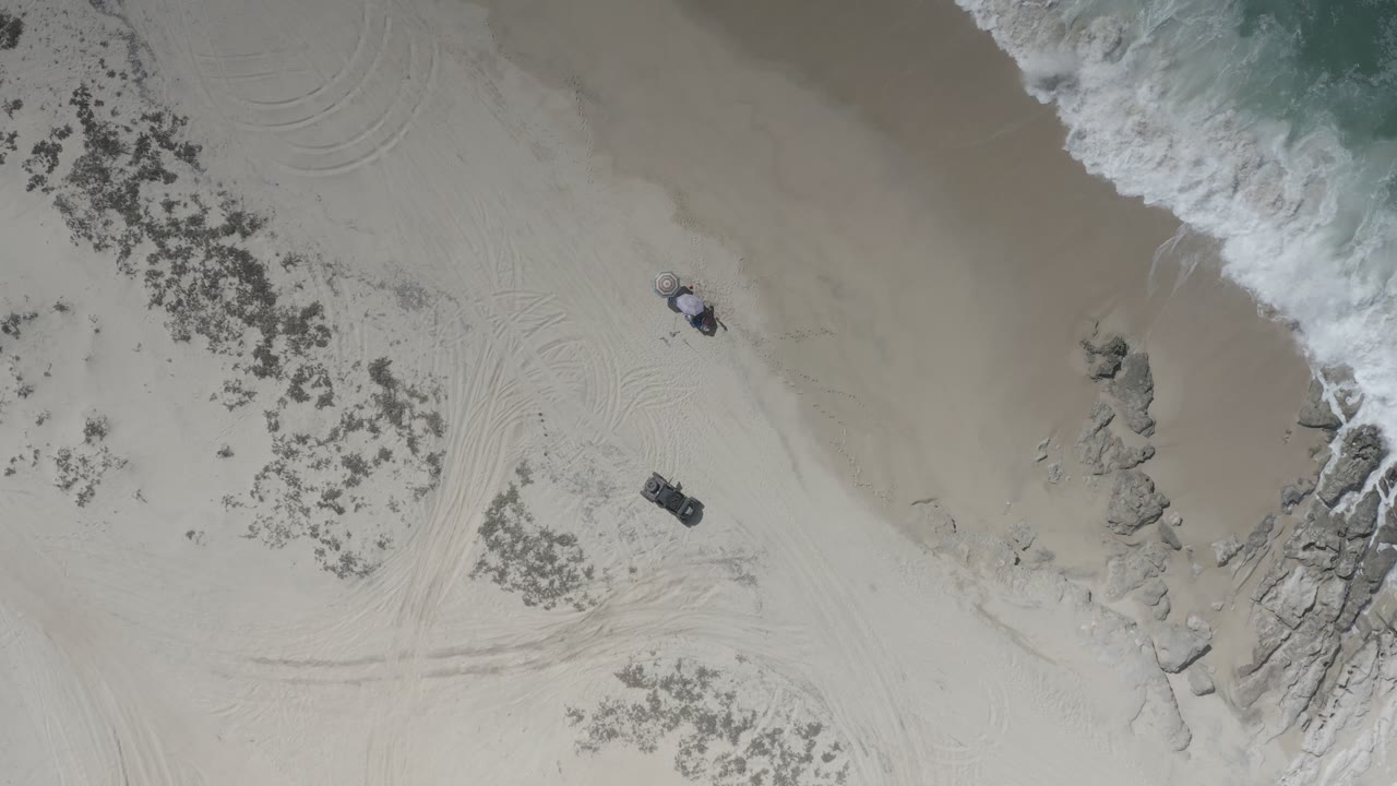 Aerial Los Cabos beach frame with the surf line pressing farther into the upper edge, two people near the open sand, rocks by the shoreline, and tire tracks curving across the beach.