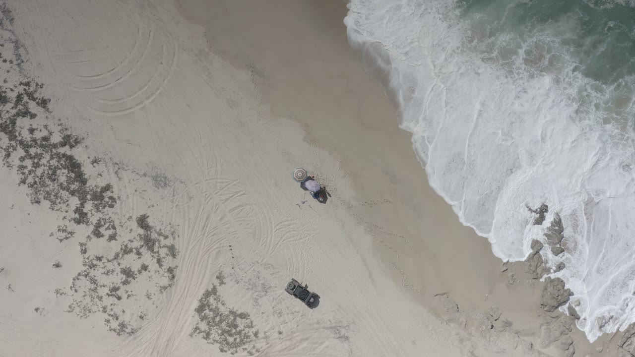 Overhead Los Cabos beach frame showing two parked vehicles on pale sand, scrub patches on the left, white surf at the upper right, and no visible people.
