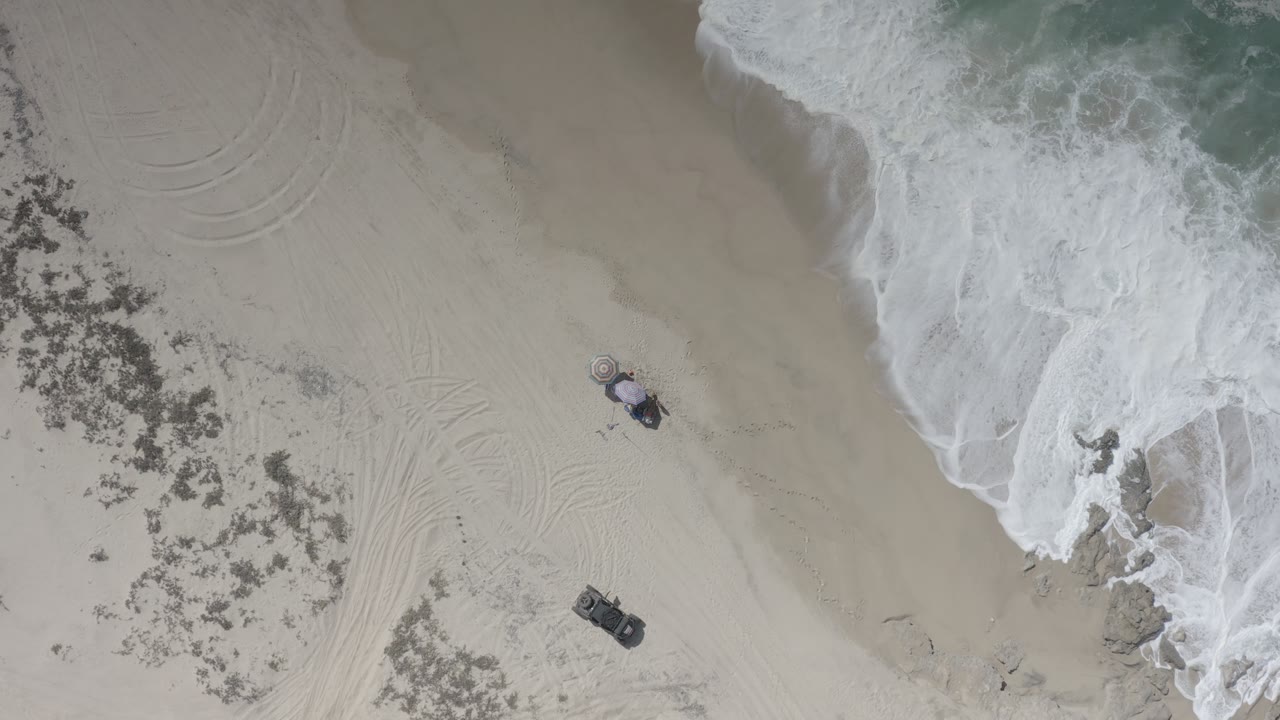 Drone view of beach tracks, parked vehicles, scrub, and foamy shoreline.