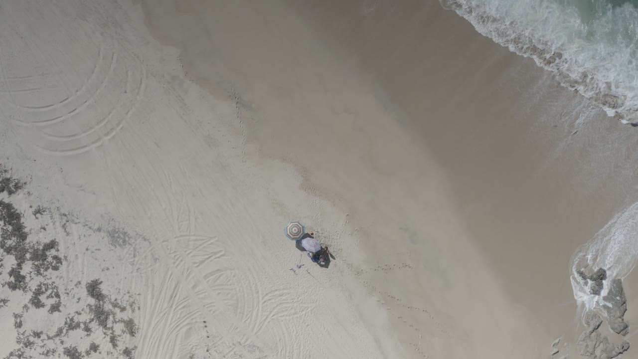 Overhead beach view with surf, sand, umbrella, and a small child.
