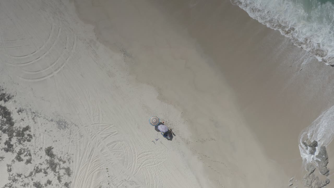 Overhead Los Cabos beach frame with the waterline running across the upper right, a small child beside an umbrella, faint curved marks in the sand, and a pale rock or foamy patch near midframe.