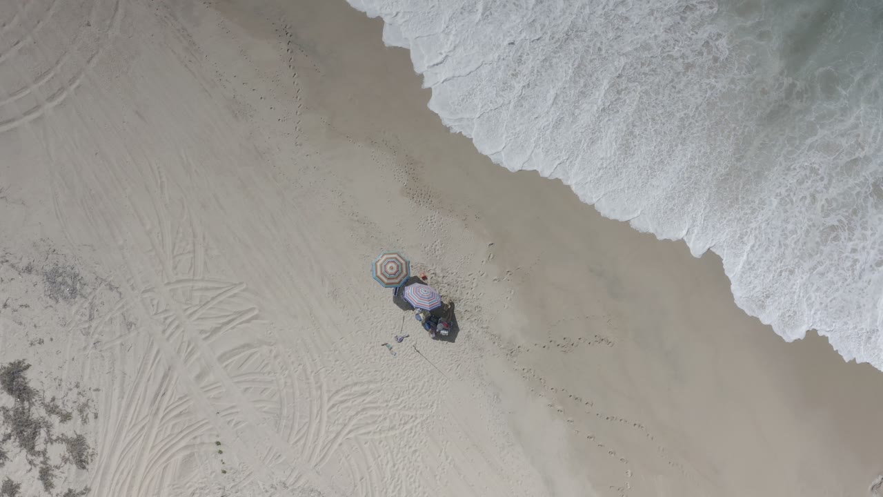 Overhead beach view with surf, sand, and a striped umbrella setup.