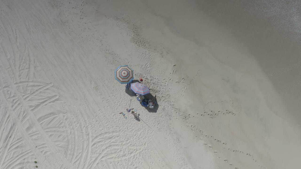 Overhead beach view with striped umbrellas, gear, footprints, and small figures on pale sand.