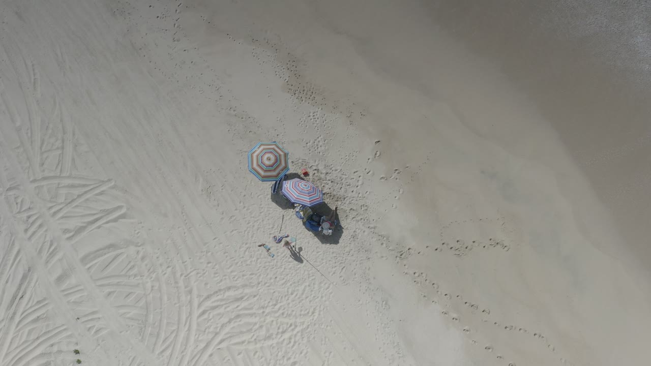 Overhead Los Cabos beach frame with two striped umbrellas, clustered beach items, faint tire tracks, footprints, and small visible figures positioned near the lower umbrella.