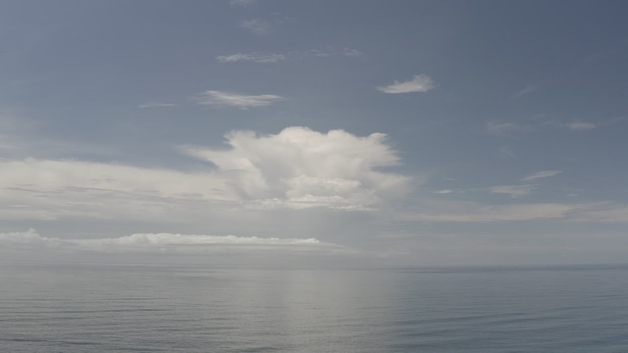 Wide Los Cabos ocean view at the start of the clip, with calm rippled water, a pale blue-gray sky, a large white cloud bank, and no visible people.