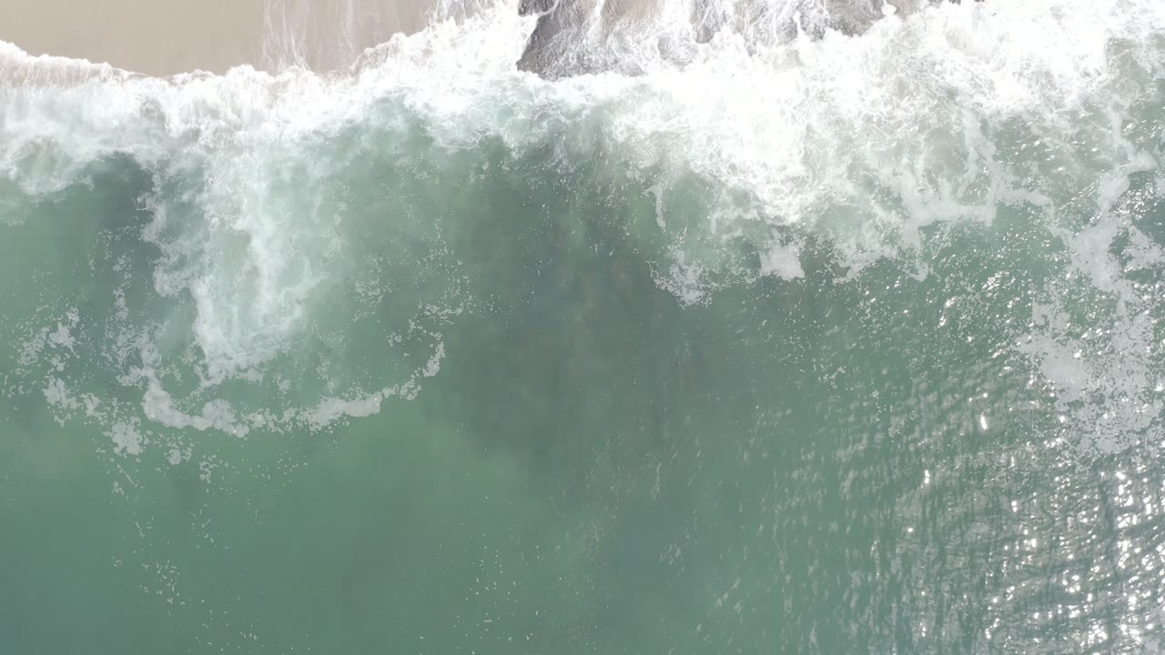 Overhead view of Los Cabos ocean surf at the start of the clip, with white foam rolling over gray-green water and no visible people.