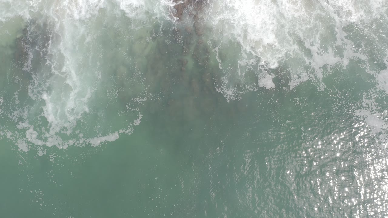 Overhead view of pale green ocean water in Los Cabos, Mexico, with white surf crossing the top of frame and no visible people.