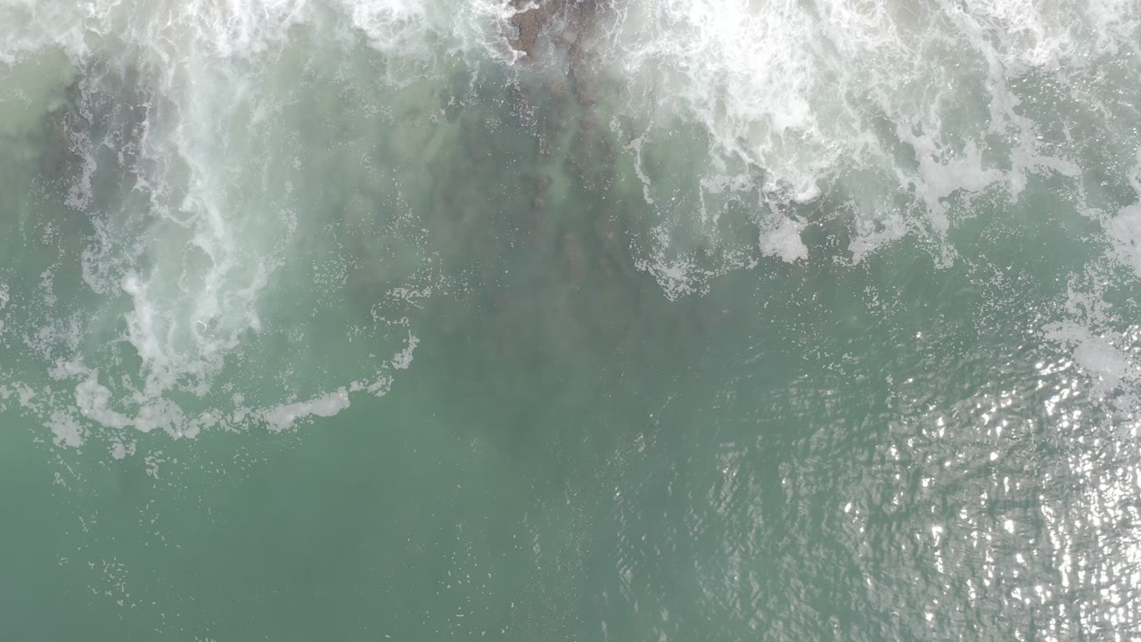 White foam spreads farther across pale green water in Los Cabos, Mexico, with bright reflected sunlight near the lower center and no visible people.