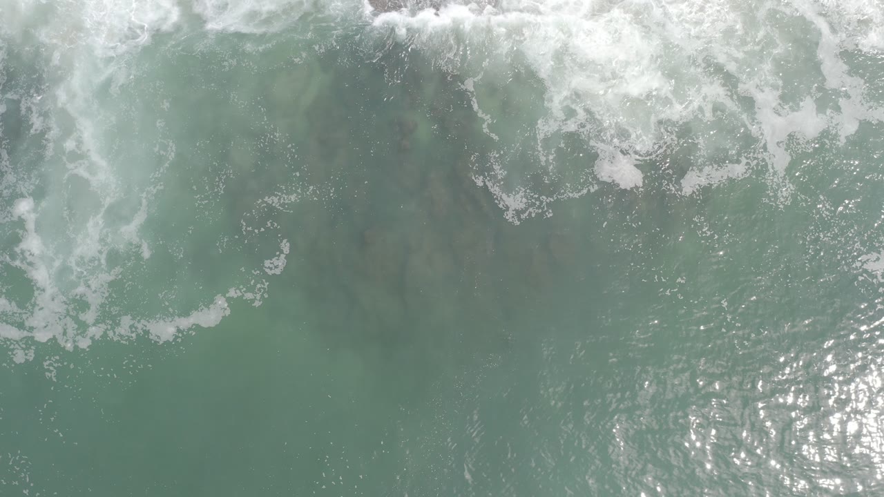 Overhead view of teal ocean water in Los Cabos, Mexico, with white surf foam across the upper frame, bright surface glints below, and no visible people.