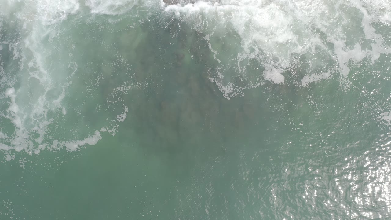 Aerial view of Los Cabos ocean water with broken white foam near the top, darker green patches under the surface, silver reflections at lower center, and no visible people.