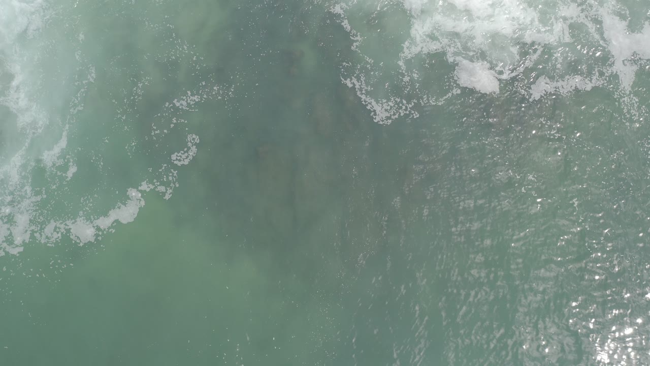 Top-down view of green-gray ocean water in Los Cabos, Mexico, with white foam drifting across the upper edge and no visible people.