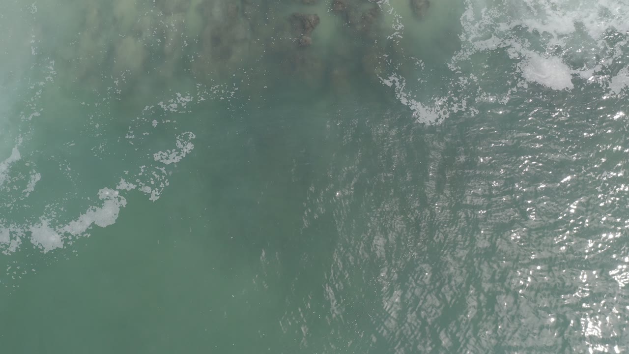 Top-down view of green-blue ocean water in Los Cabos, Mexico, with scattered foam and no visible people at the start of the clip.
