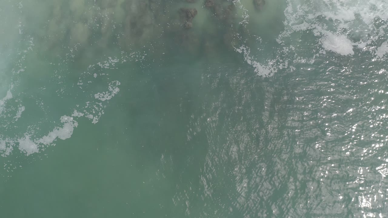 Overhead Los Cabos ocean water shows brighter sun reflection across ripples, white foam near the top, and no visible people.