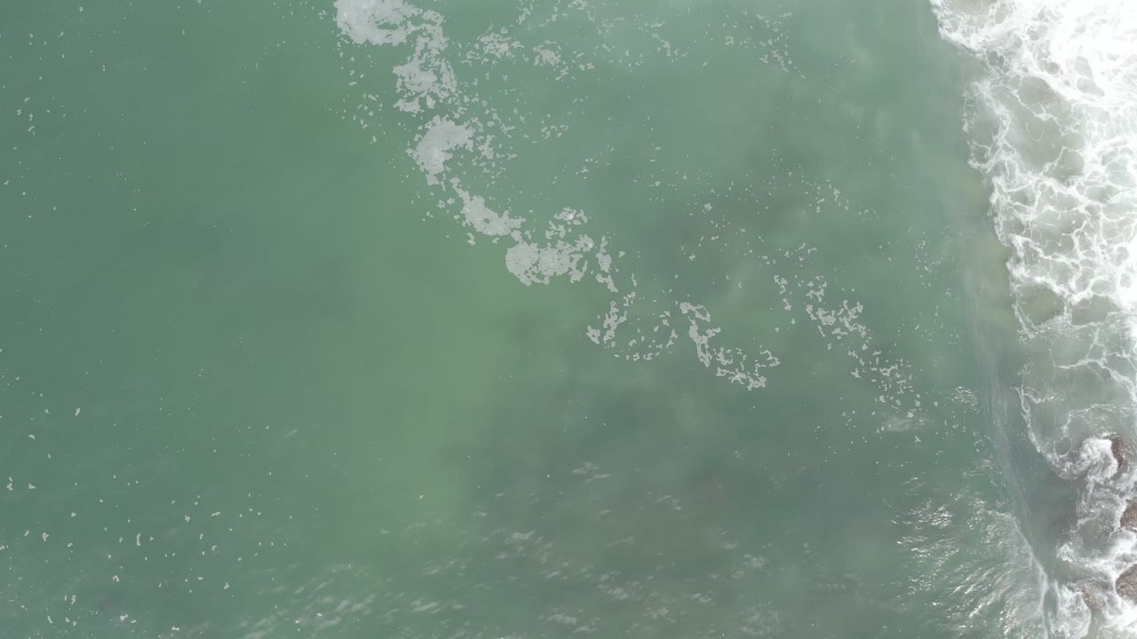 Overhead aerial view in Los Cabos, Mexico showing green ocean water, scattered white foam, and a breaking surf line near the right edge with no visible people.