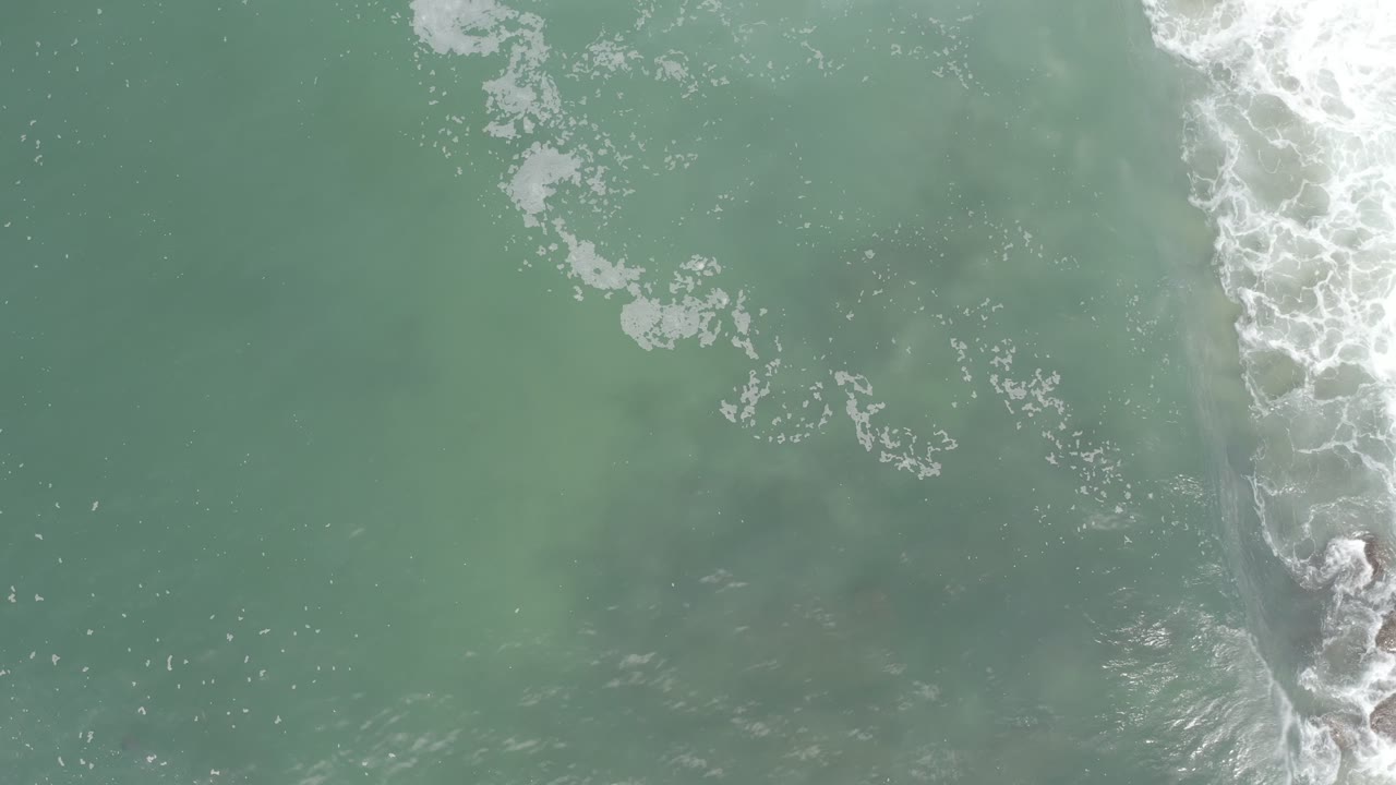Overhead aerial frame from Los Cabos, Mexico with pale green sea, a vertical band of white wave foam, and darker underwater areas, with no visible people.
