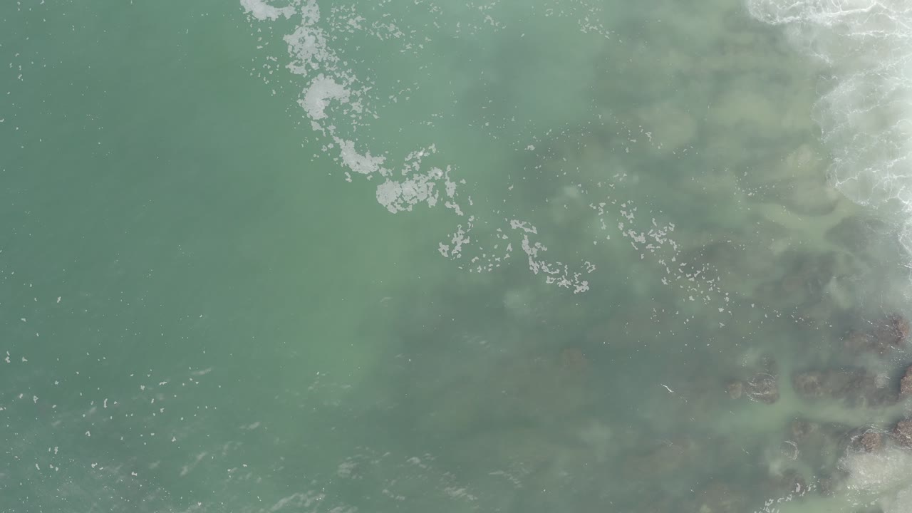 Overhead view of green-blue ocean water in Los Cabos, Mexico, with white foam entering from the upper right and no visible people.