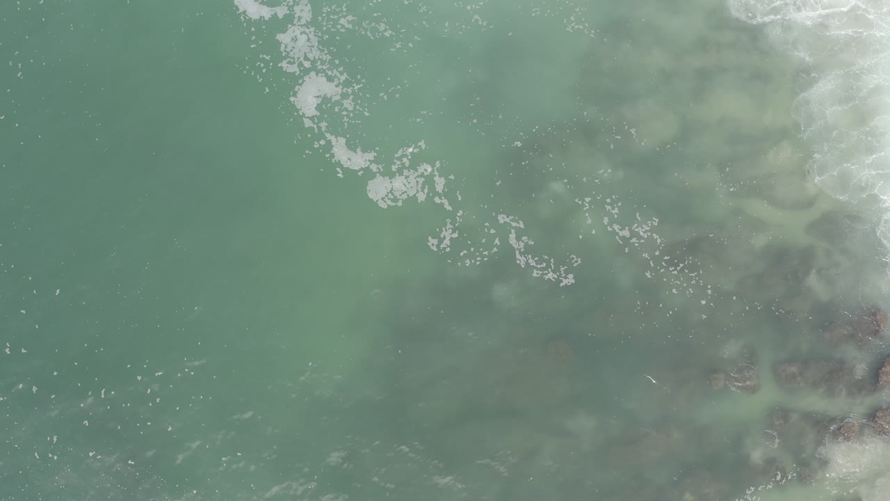 Aerial overhead frame of Los Cabos ocean water, with scattered white foam near the top and darker underwater forms at the lower right, no visible people.