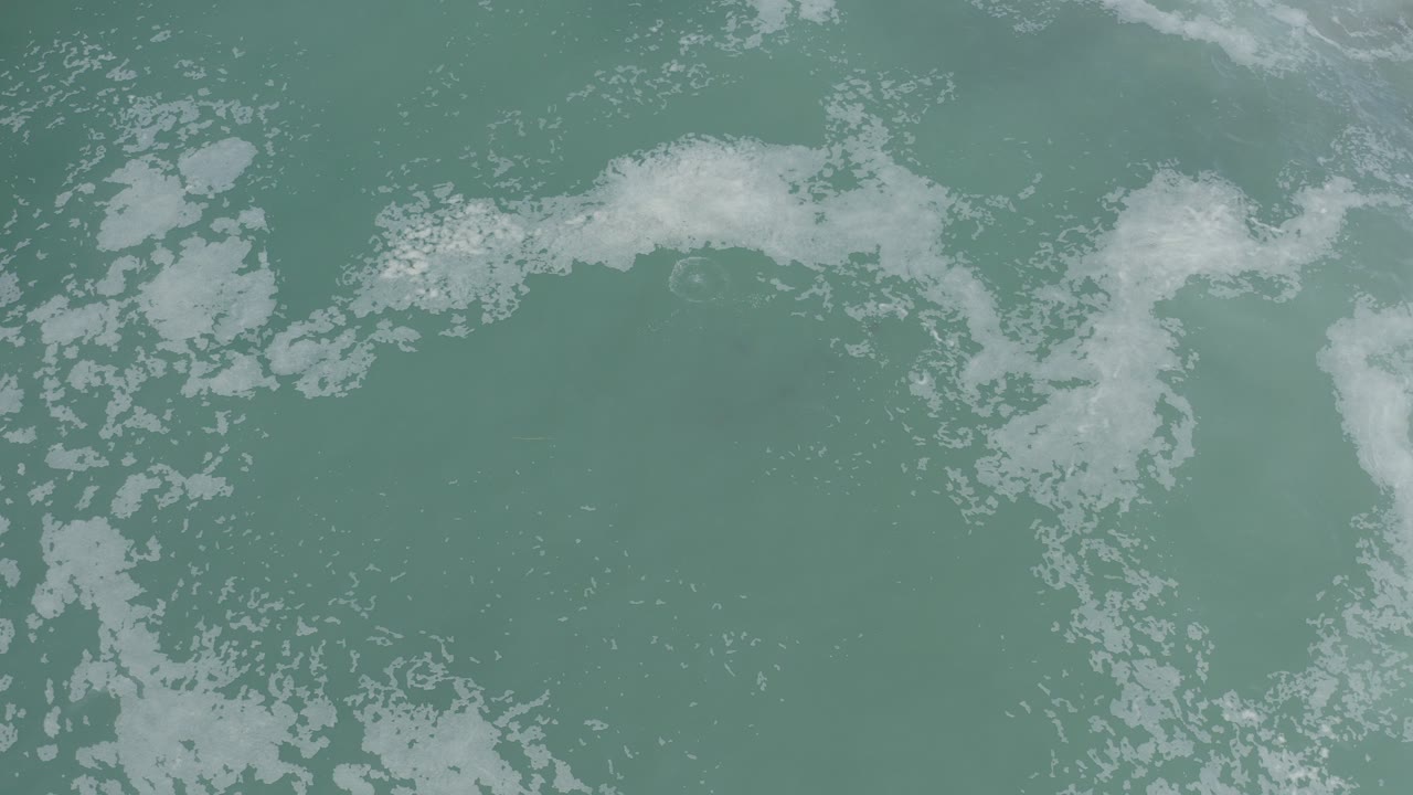 White foam patches drift across cloudy turquoise-green ocean water in Los Cabos, Mexico, with no visible people.