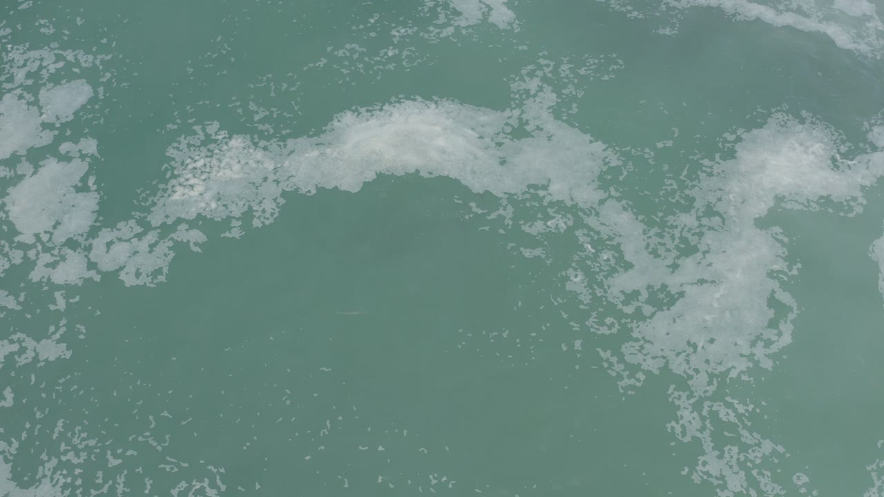 Overhead view of blue-green seawater in Los Cabos with a broken band of white foam near the top of frame and no visible people.