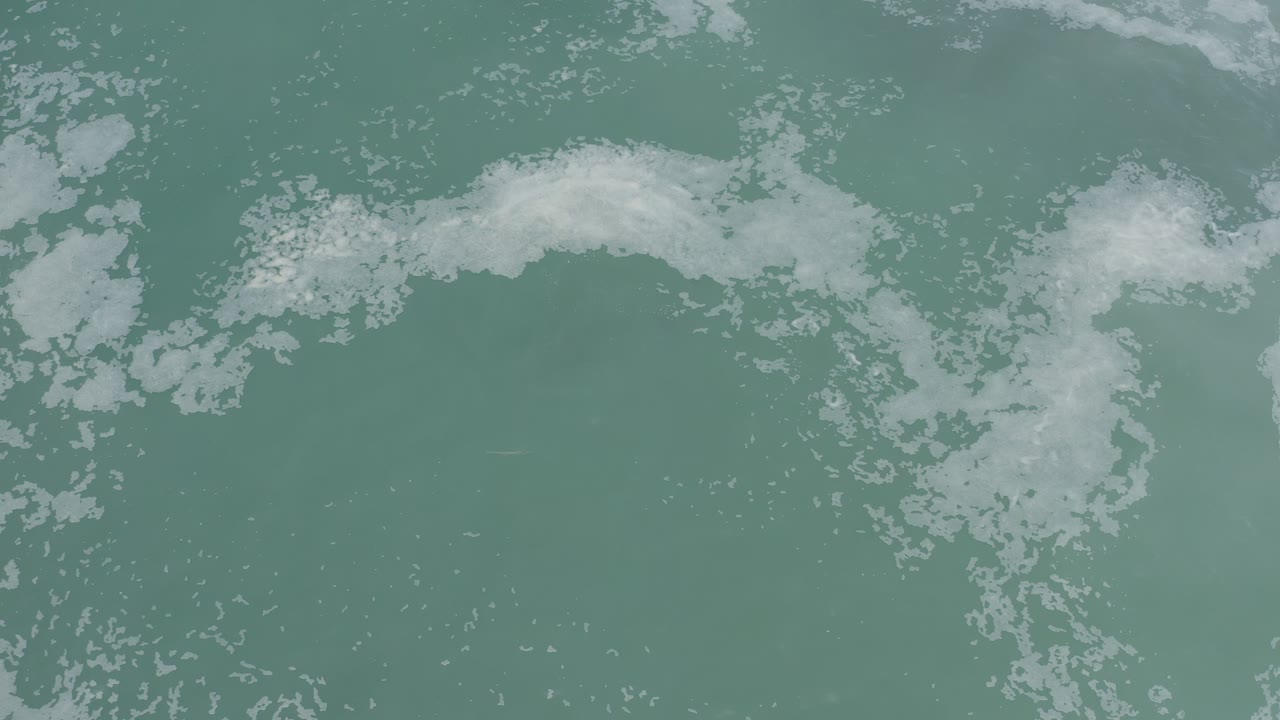 Los Cabos ocean surface seen from above, with a curved white foam patch crossing the upper half and fine bubbles below, no visible people.