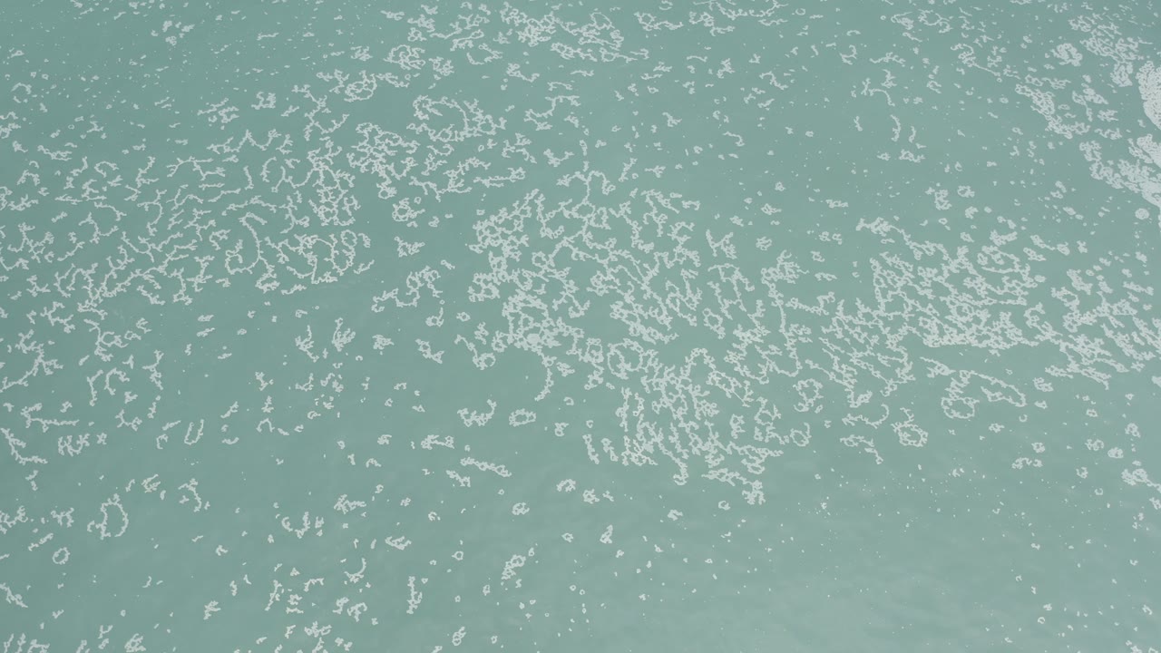 Overhead view of pale turquoise water in Los Cabos, Mexico, with white foam patterns across the upper half and no visible people.