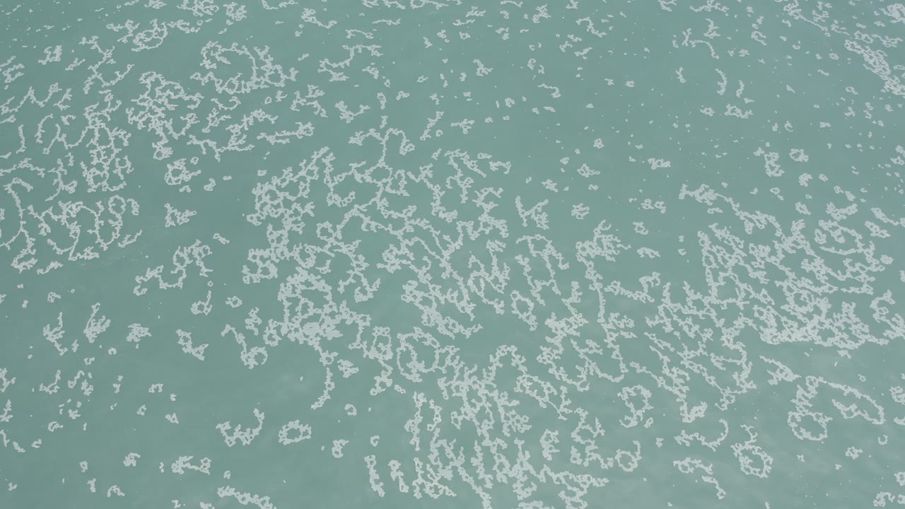 Overhead view of pale turquoise ocean water in Los Cabos, Mexico, with scattered white sea foam patterns and no visible people.