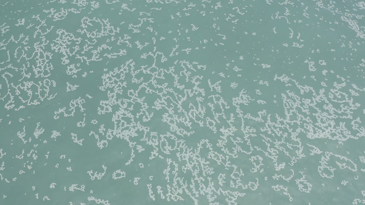 A later overhead frame shows thin white foam streaks spread across muted turquoise water in Los Cabos, Mexico, with no visible people.