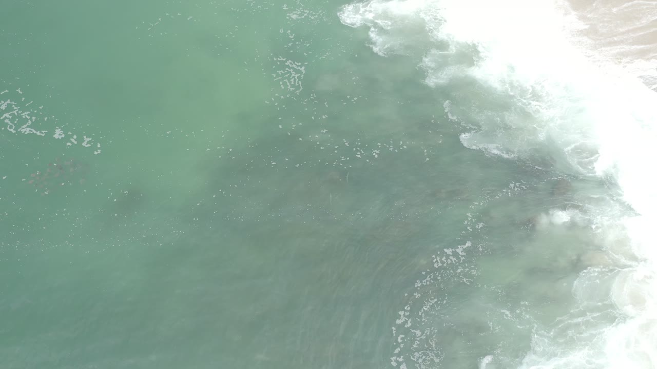 Overhead view of blue-green ocean water over sandy shallows in Los Cabos, Mexico, with bright white foam along the right side and no visible people.