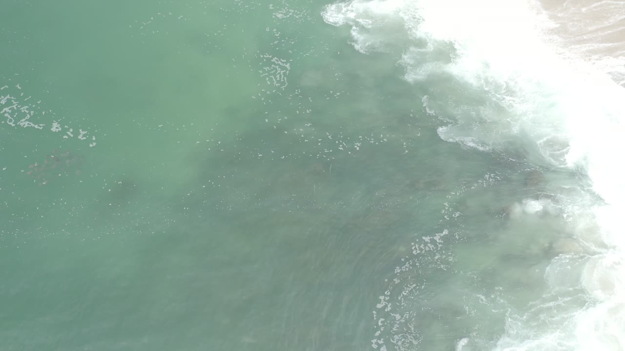 Overhead Los Cabos ocean frame with white foam spreading from the upper right across blue-green water, sandy bottom visible beneath, and no visible people.