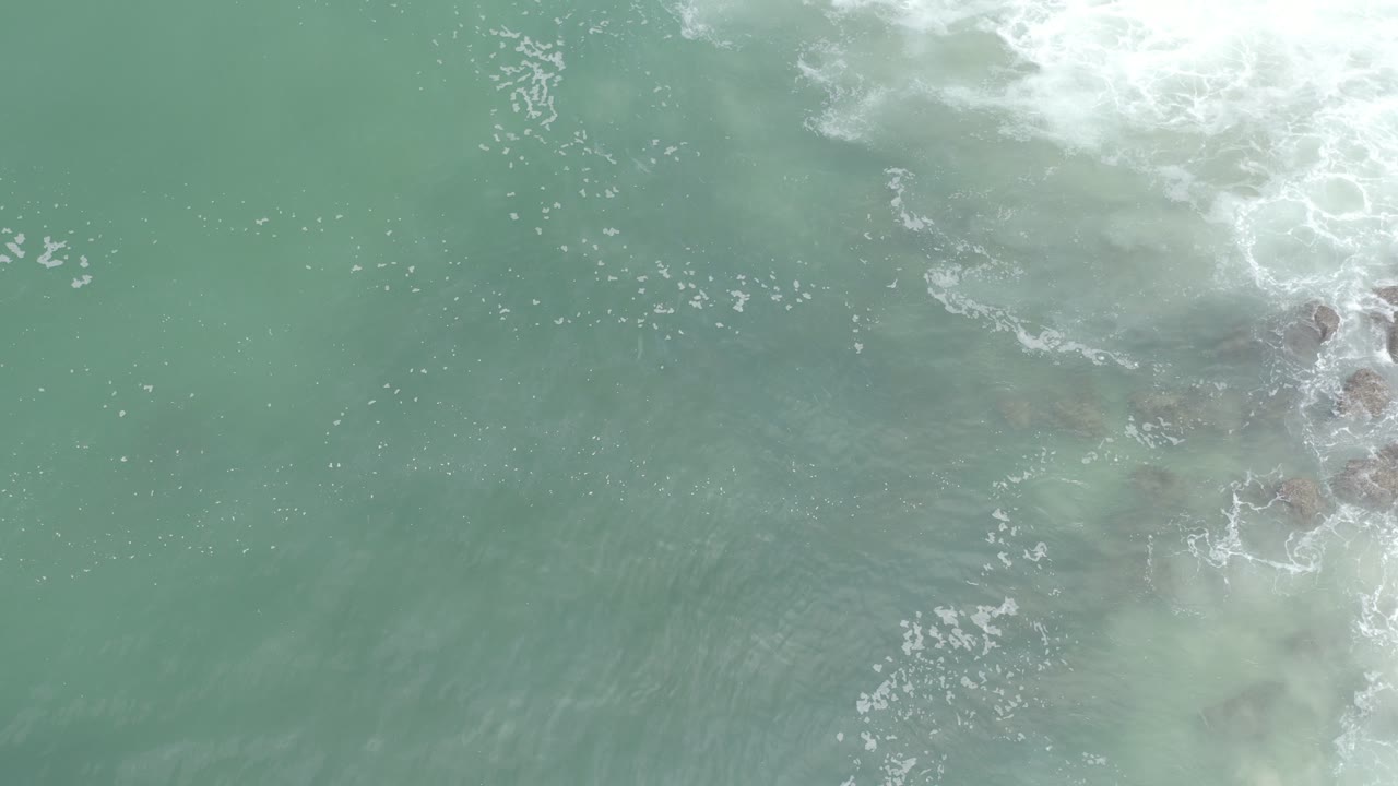 Overhead view of grayish blue-green ocean in Los Cabos, Mexico, with white surf foam along the upper edge and no visible people.