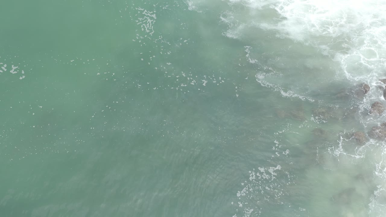 Overhead Los Cabos water frame with pale green-blue surface, frothy white wash at the upper right, and no visible people.