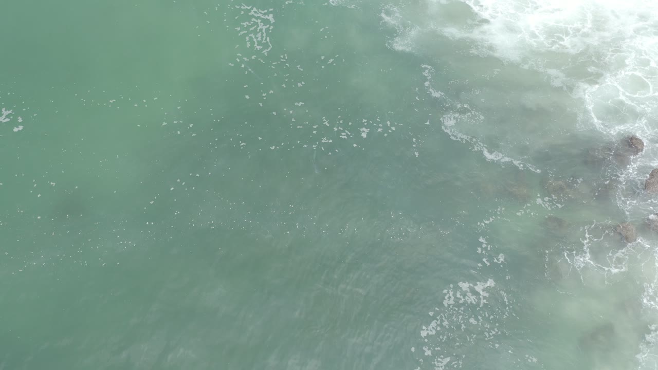 Aerial view of muted turquoise water with foam and darker shapes near the right edge.