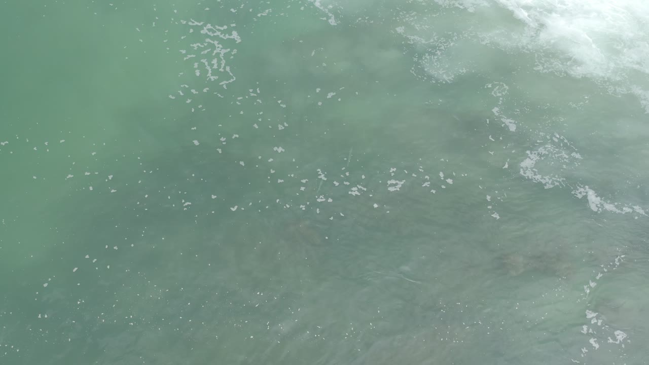 Overhead Los Cabos ocean frame with muted green water, small white foam flecks, darker submerged areas, and no visible people.