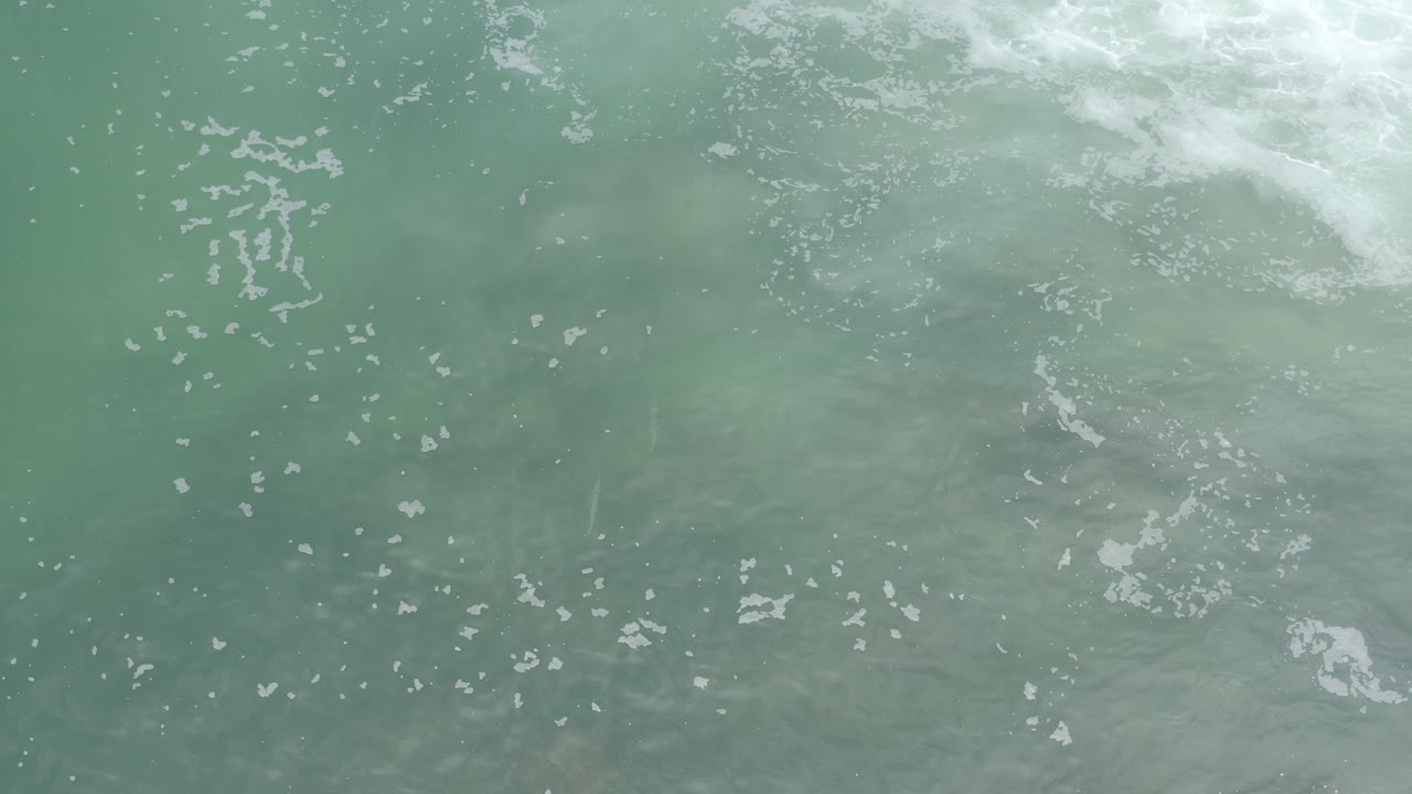 Overhead view of green-blue seawater in Los Cabos, Mexico, with scattered pale foam near the top edge and no visible people.