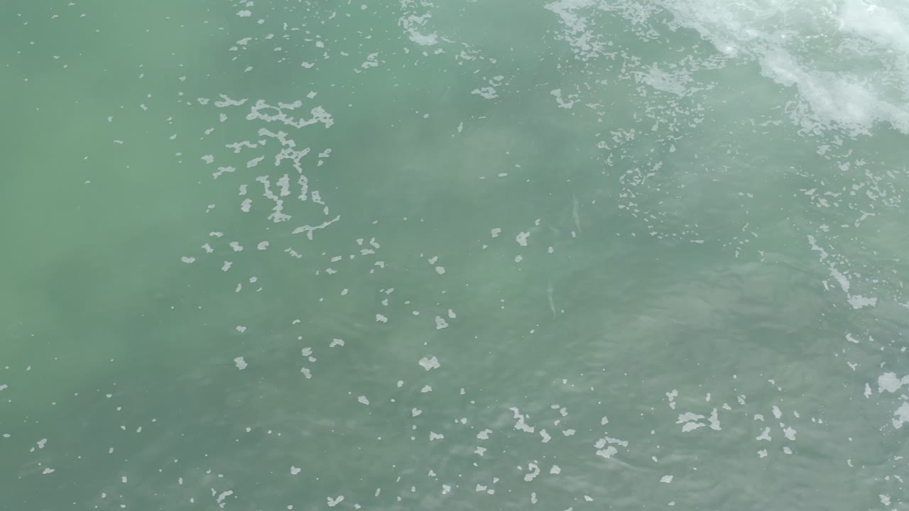 Aerial view over Los Cabos ocean water where small white foam flecks sit above faint darker underwater patches, with no visible people.