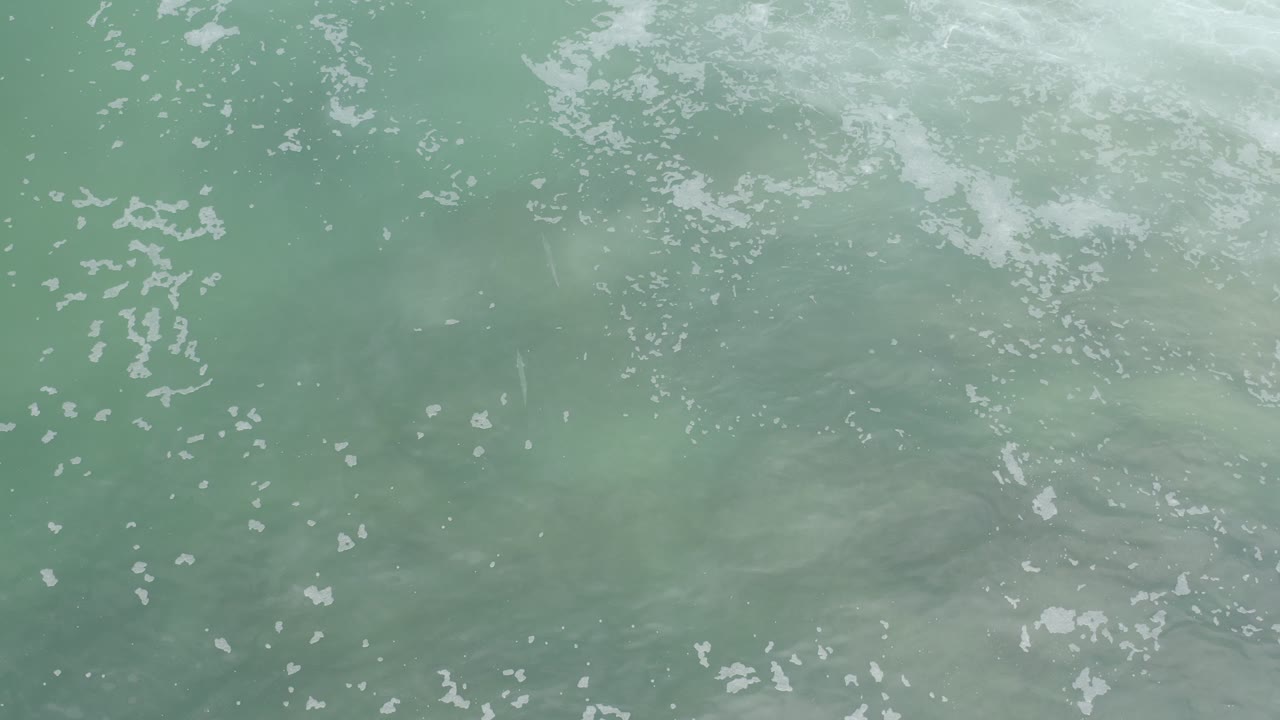 Overhead view of pale turquoise water in Los Cabos with small white foam patches near the upper edge and no visible people.