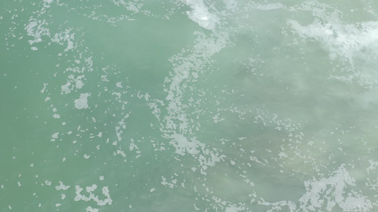 Overhead view of pale green sea water in Los Cabos with small white foam clusters spread across the surface and no visible people.