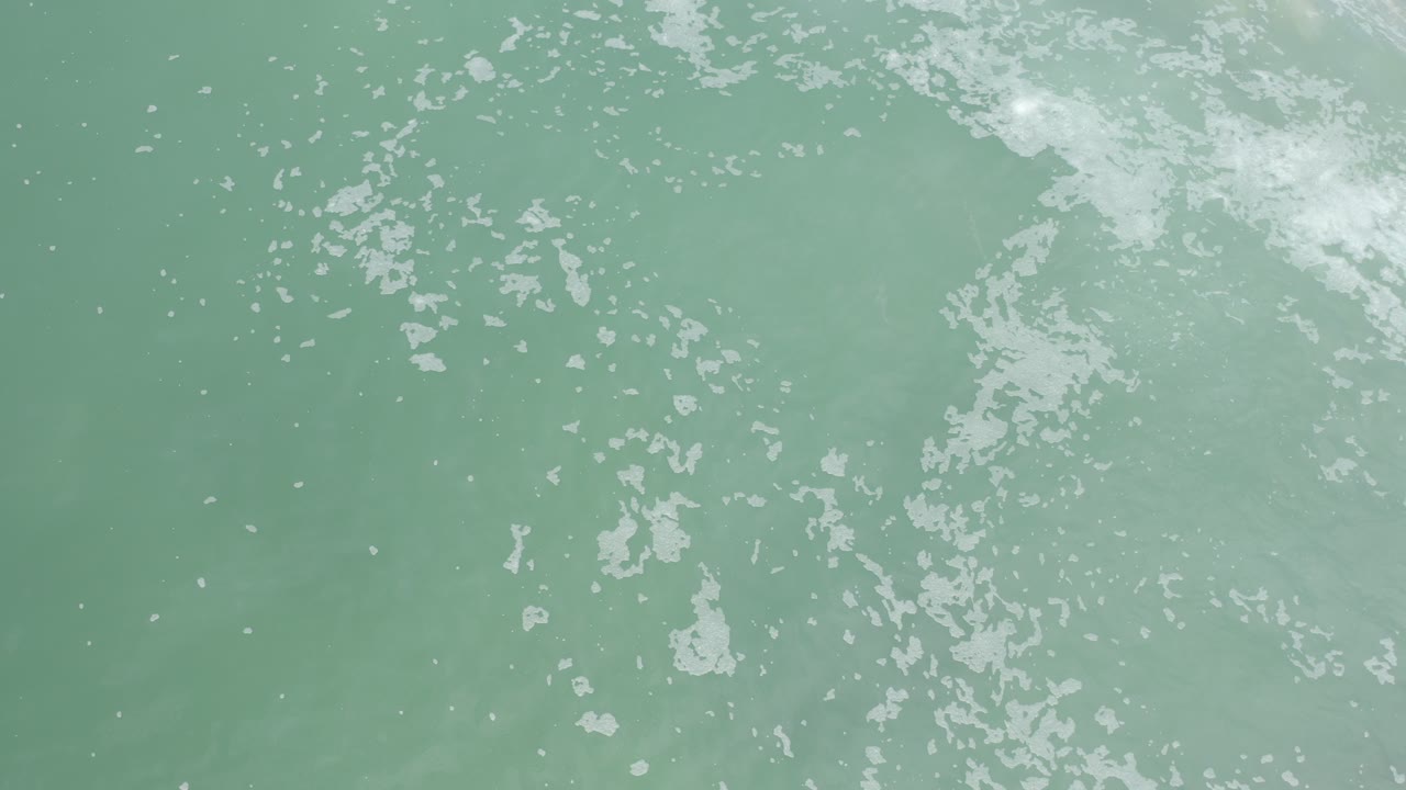 Overhead view of pale green ocean water in Los Cabos with white surf foam clustered near the upper edge and no visible people.